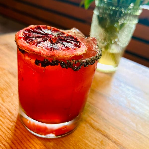 A vibrant red cocktail with a charred orange slice and dark rim, on a wooden table.