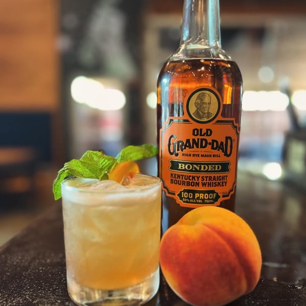 A bourbon cocktail with mint and peach, next to a bottle of Old Grand-Dad bourbon and a fresh peach.