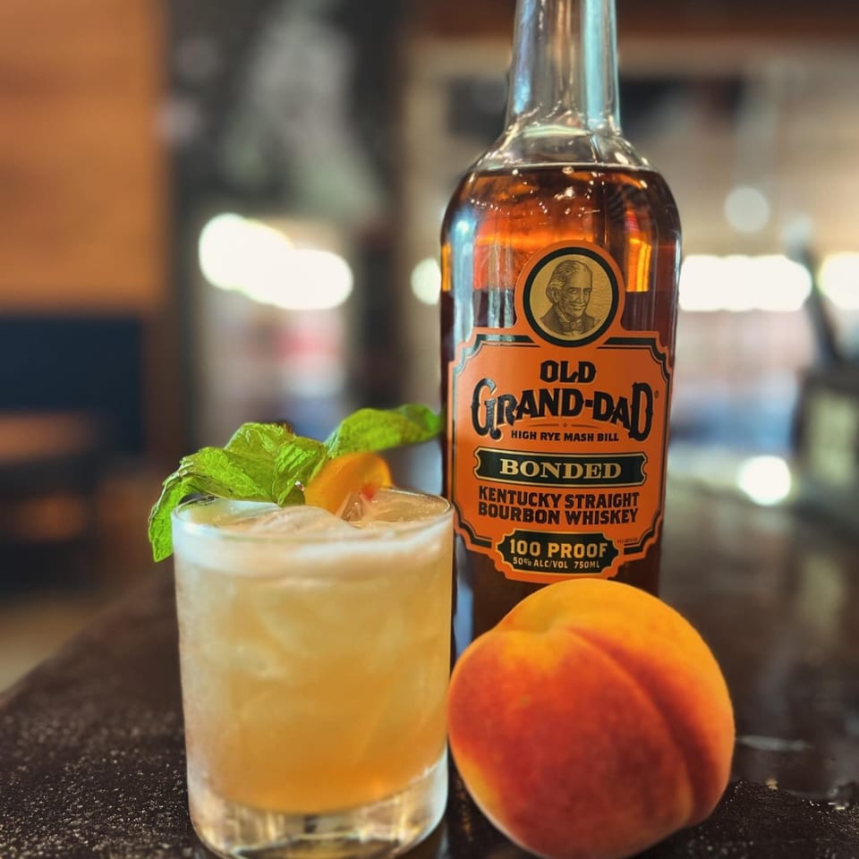 A bourbon cocktail with mint and peach, next to a bottle of Old Grand-Dad bourbon and a fresh peach.